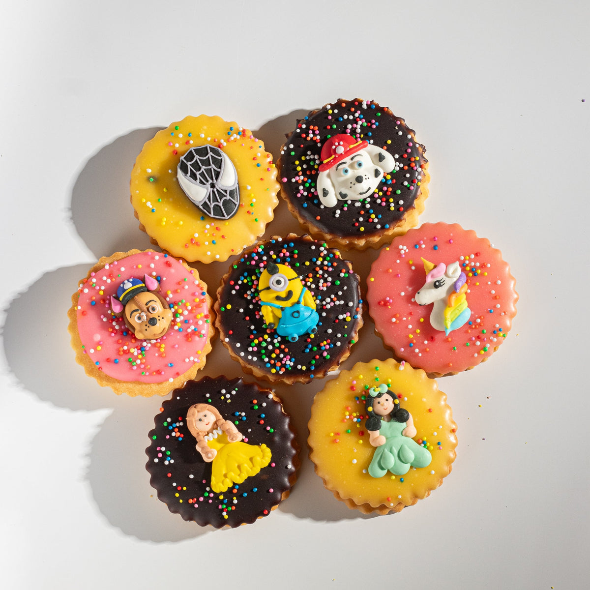Seven decorated cupcakes arranged in a loose circle on a white background, each topped with colorful sprinkles and character or animal figurines including Spider-Man, Dalmatian, monkey, cartoon character, unicorn, baby, and princess.