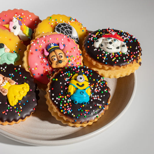 A group of round iced cookies decorated with colorful sprinkles and character toppers, arranged on a light beige plate against a clean white background.