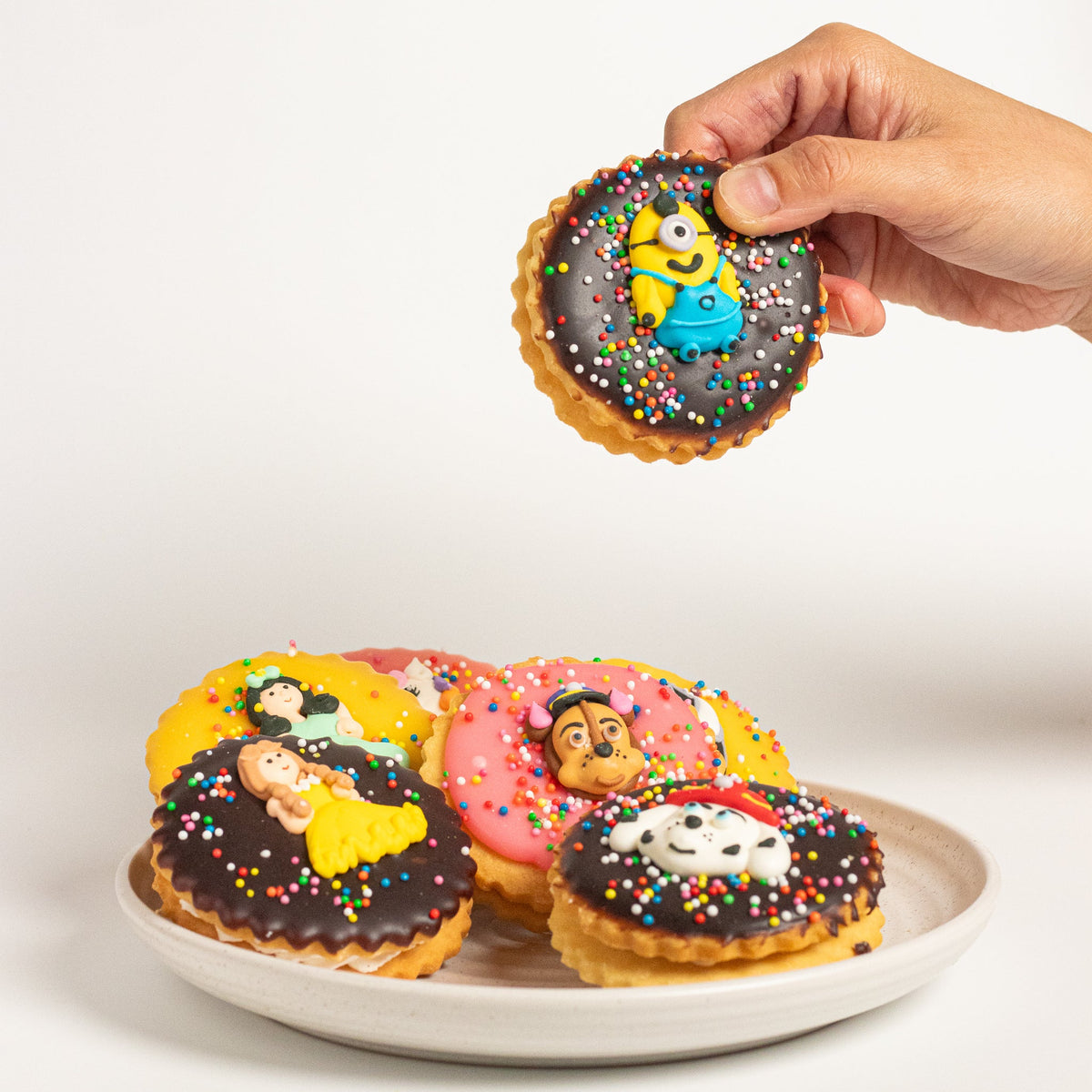 A hand holds a round iced cookie featuring a Minions character above a white plate of several colorful character cookies on a white background.
